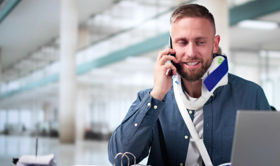 A man wearing a neck brace at an office talks on his phone while looking at his laptop.