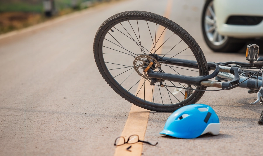 A bicycle, a helmet, and a pair of damaged glasses lay on the road in front of a parked car.