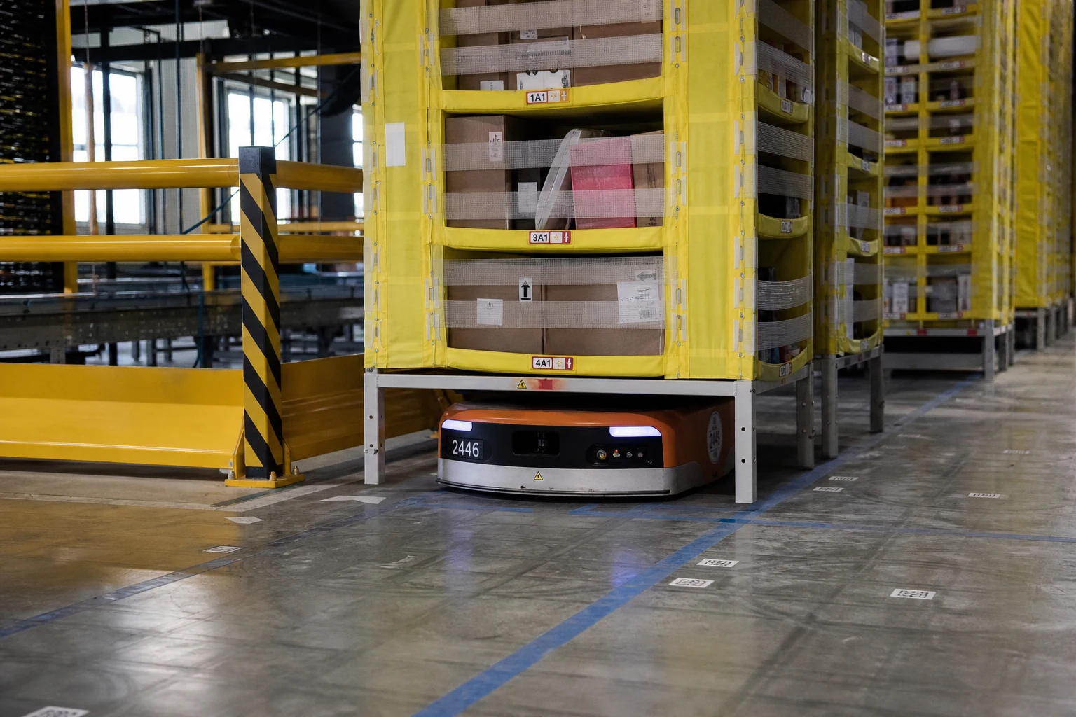 Low orange autonomous mobile shelf-carrying robot under a tall industrial shelving pod inside a warehouse with yellow caution chevrons on the surrounding guard rail — the kind of robotics equipment a Fort Wayne Amazon warehouse injury attorney pursues as a third-party defendant