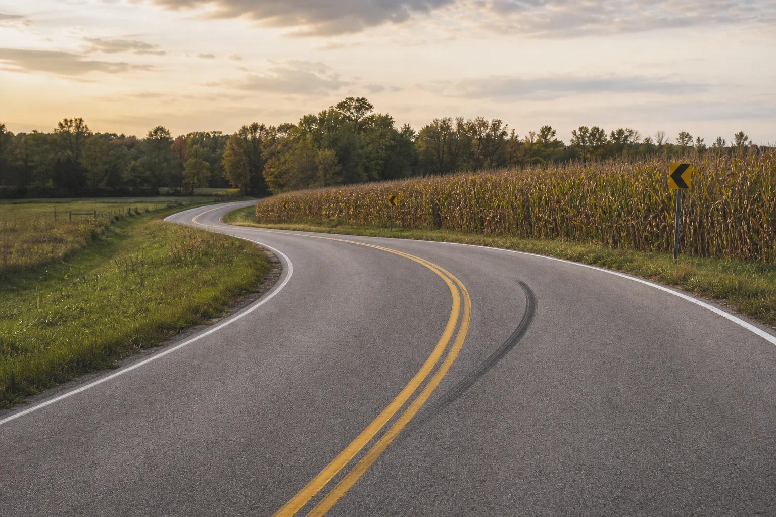Indiana rural two-lane road curving past a cornfield at golden hour with a single black skid mark on the asphalt — the kind of corridor where Allen County motorcycle wrecks happen most