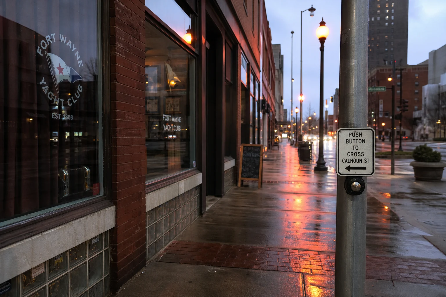 Downtown Fort Wayne sidewalk along a brick storefront block at twilight, red-orange streetlamps glowing, a pedestrian-crossing button on a steel pole, wet pavement reflecting — where most Allen County pedestrian-attorney cases originate