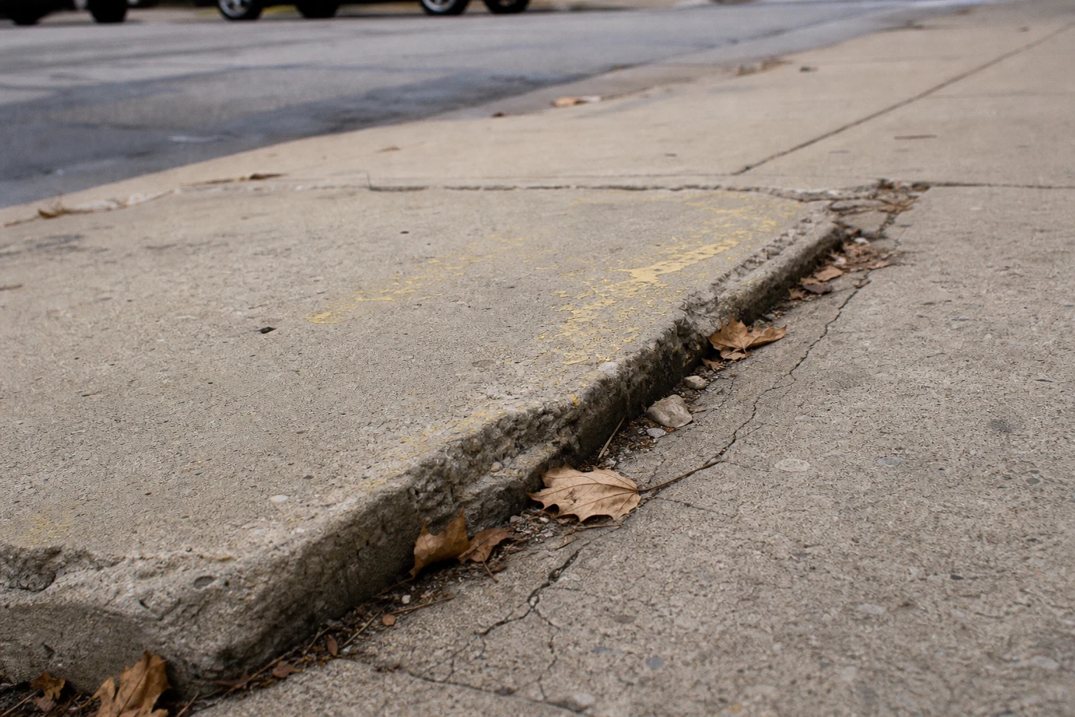 Heavily cracked Fort Wayne concrete sidewalk slab with a raised edge that could trip a pedestrian, faded yellow paint, autumn leaves in the seam — the kind of premises hazard a Fort Wayne slip trip and fall attorney documents the day the file opens