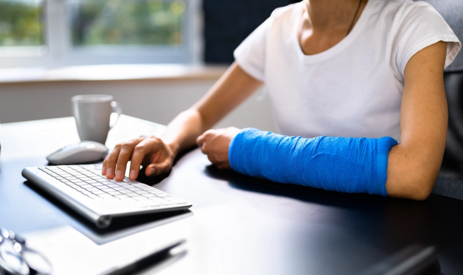 A woman types on her keyboard one-handed as the other arm sits on the desk in a cast.