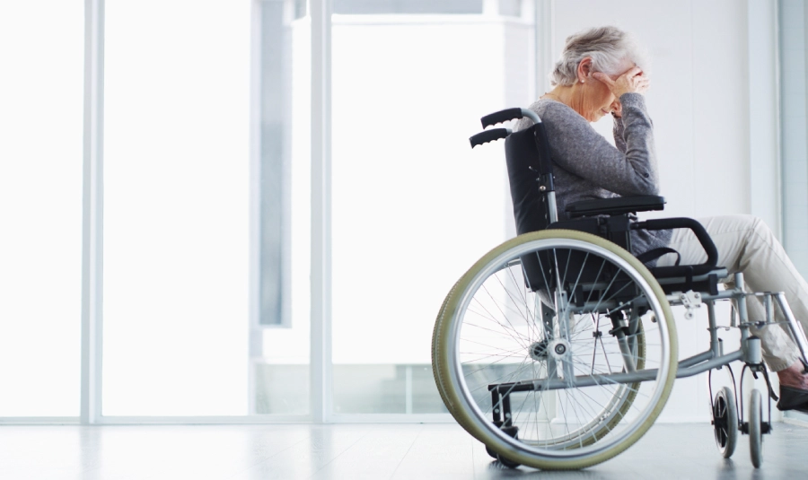 An elderly woman holder her face in her hands while sitting in a wheelchair.